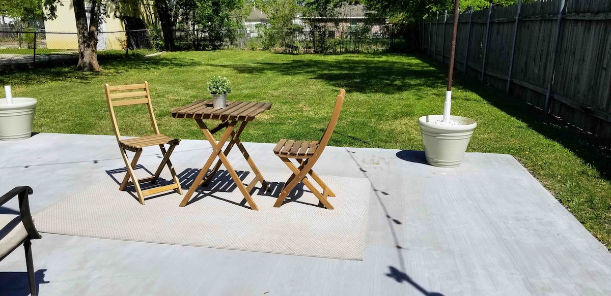 A patio area features a small wooden table accompanied by two folding chairs, set on a stone surface. The surrounding green grass is complemented by planters with foliage. A rug lies beneath the table, and a tree provides shade in the background.
