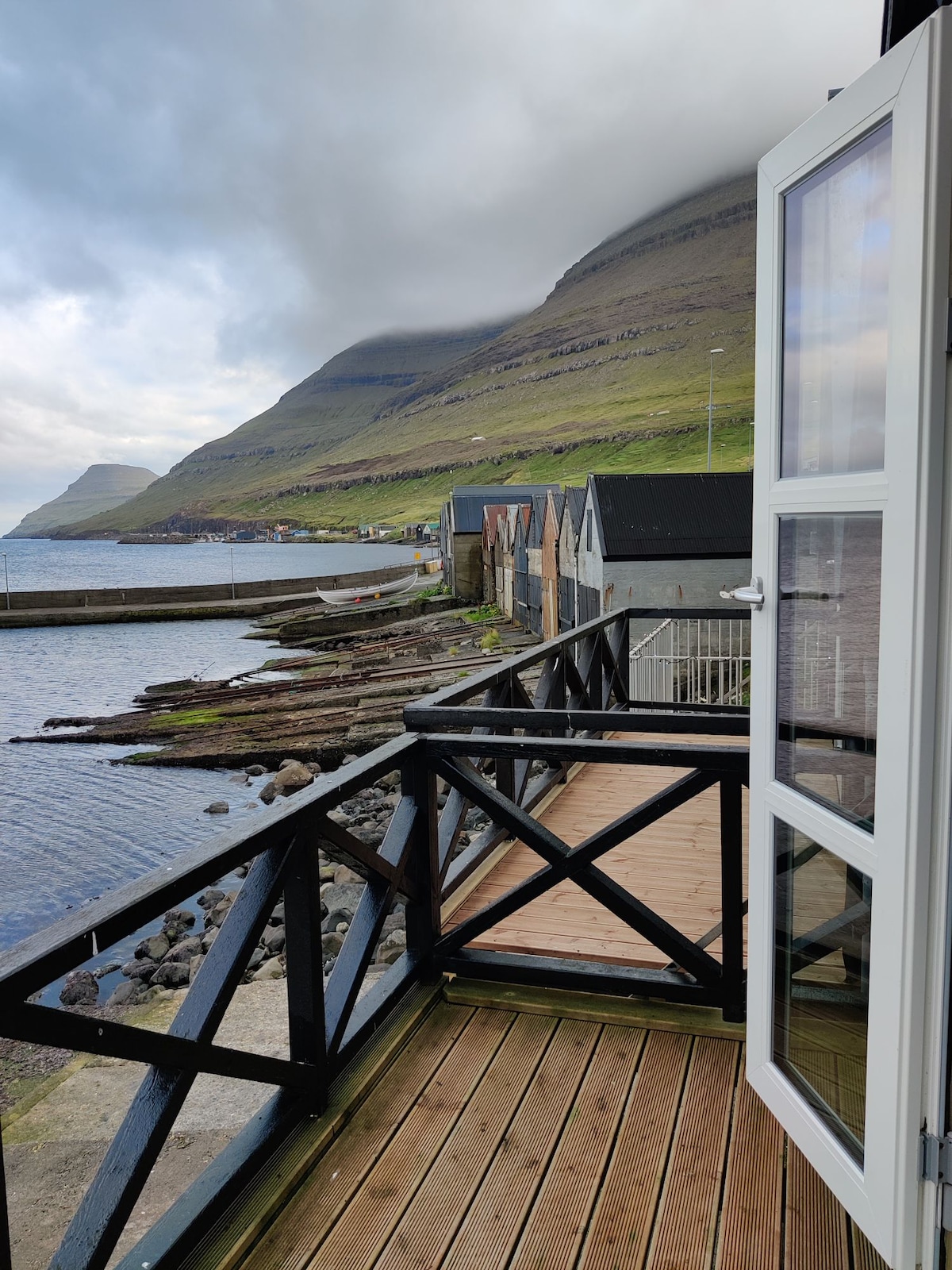 A wooden balcony with a black railing overlooks a rocky shore and a tranquil sea. The mountain rises in the background, partially obscured by clouds. Nearby, traditional buildings can be seen alongside the water's edge, offering scenic views of the serene landscape.
