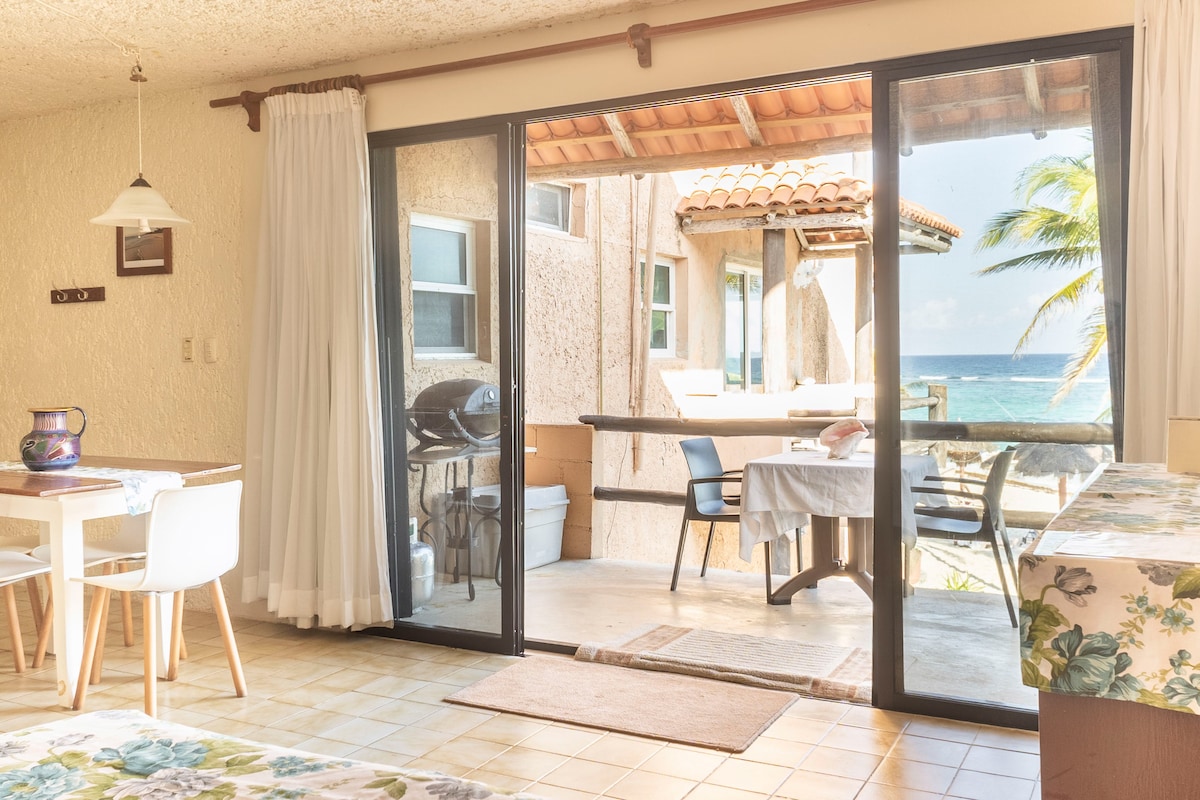 A welcoming indoor space features a dining area with a table and white chairs, and a cozy sleeping area with a patterned bedspread. Large glass doors open to a view of the ocean, allowing natural light to fill the room.