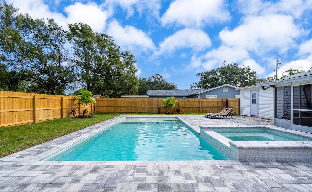 A spacious backyard features a newly renovated heated pool and hot tub, surrounded by a brick patio. Lush green grass provides ample space for outdoor activities, while a wooden fence offers privacy. Clear blue skies and scattered clouds enhance the serene atmosphere.