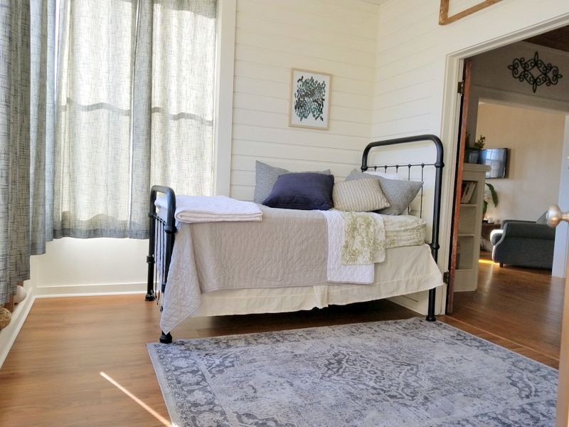 A sunroom features a daybed adorned with various textured pillows and blankets. Natural light filters through sheer, light-colored curtains, illuminating the wood flooring. A patterned area rug adds a touch of comfort, while a doorway leads to the adjoining living area.