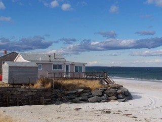 A sandy beach is visible in the foreground, leading towards a cottage featuring wrap-around windows. The house has a deck that overlooks the ocean, where gentle waves can be seen under a partly cloudy sky. The surrounding landscape includes patches of grass and rocky formations.