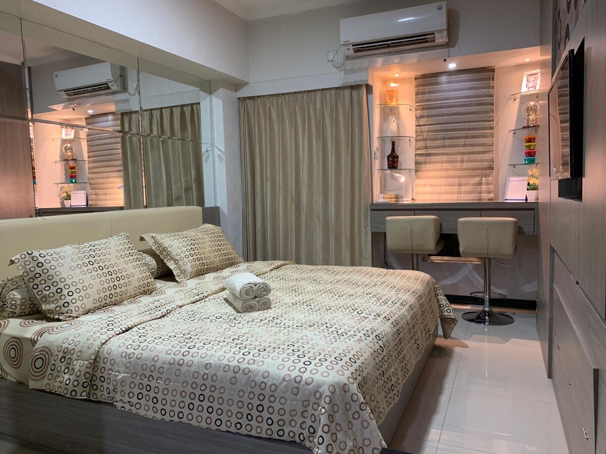 The studio features a spacious bed covered in a beige patterned linen set, accompanied by a rolled towel. Mirrored walls enhance the sense of space, while a work area with two stools is positioned beside a window with soft curtains.