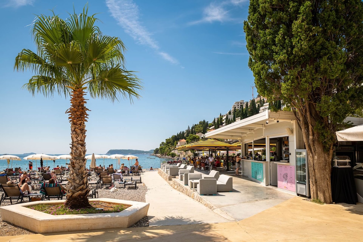 A beachfront area is depicted with a clear blue sky and gentle waves. Umbrellas and lounge chairs are arranged along the sandy shore, while a casual seaside bar is visible, surrounded by palm trees. People enjoy the sun and relax near the water.