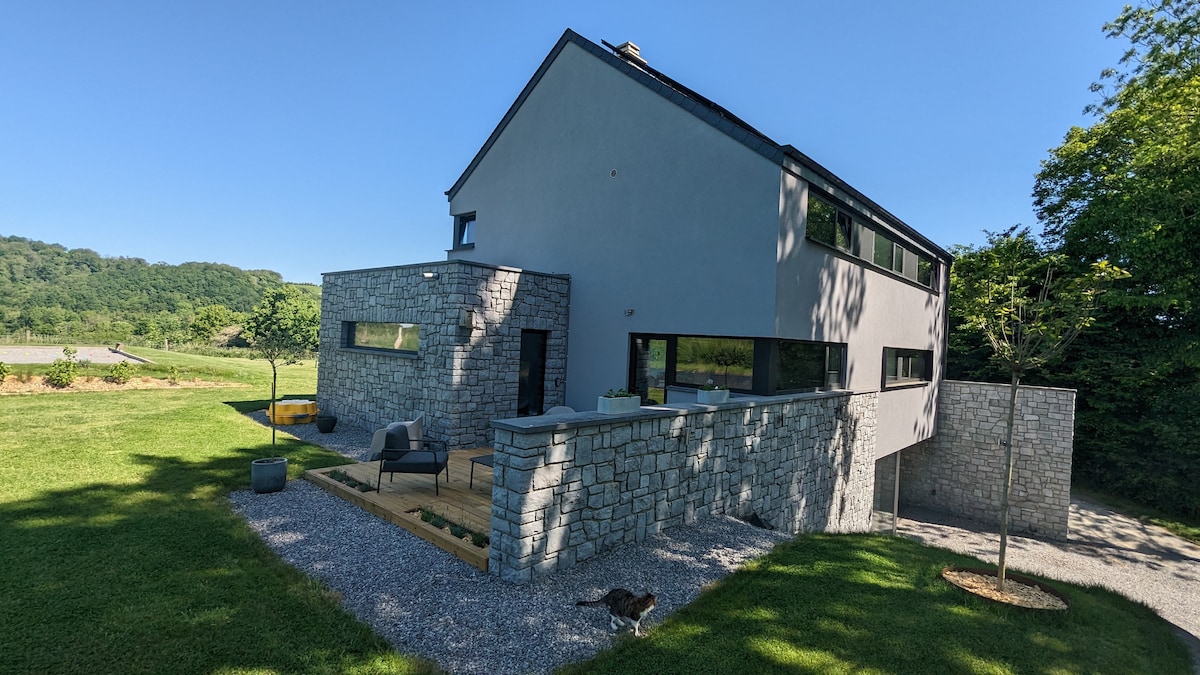 A modern villa features a stone exterior with large windows, set within a green landscape. The outdoor area includes a wooden terrace with seating, surrounded by a manicured lawn and trees. Nearby, a gravel path leads to a tranquil countryside view.