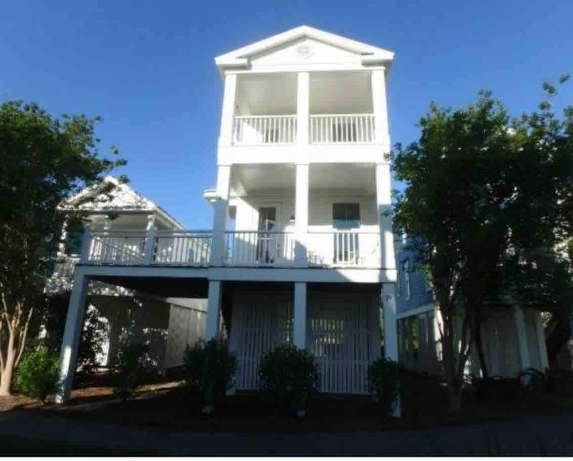 A three-story house is depicted with a classic white exterior and a covered porch. Balconies on each level are accessible by white railings. Lush greenery surrounds the base, contributing to a welcoming façade.