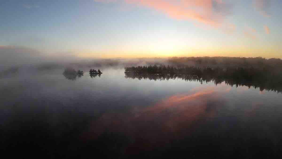 A serene lake scene at sunrise is captured, with soft pastel colors reflecting on the water's surface. Mist hovers over the lake, and silhouettes of tree lines can be seen in the background, creating a peaceful and tranquil atmosphere.