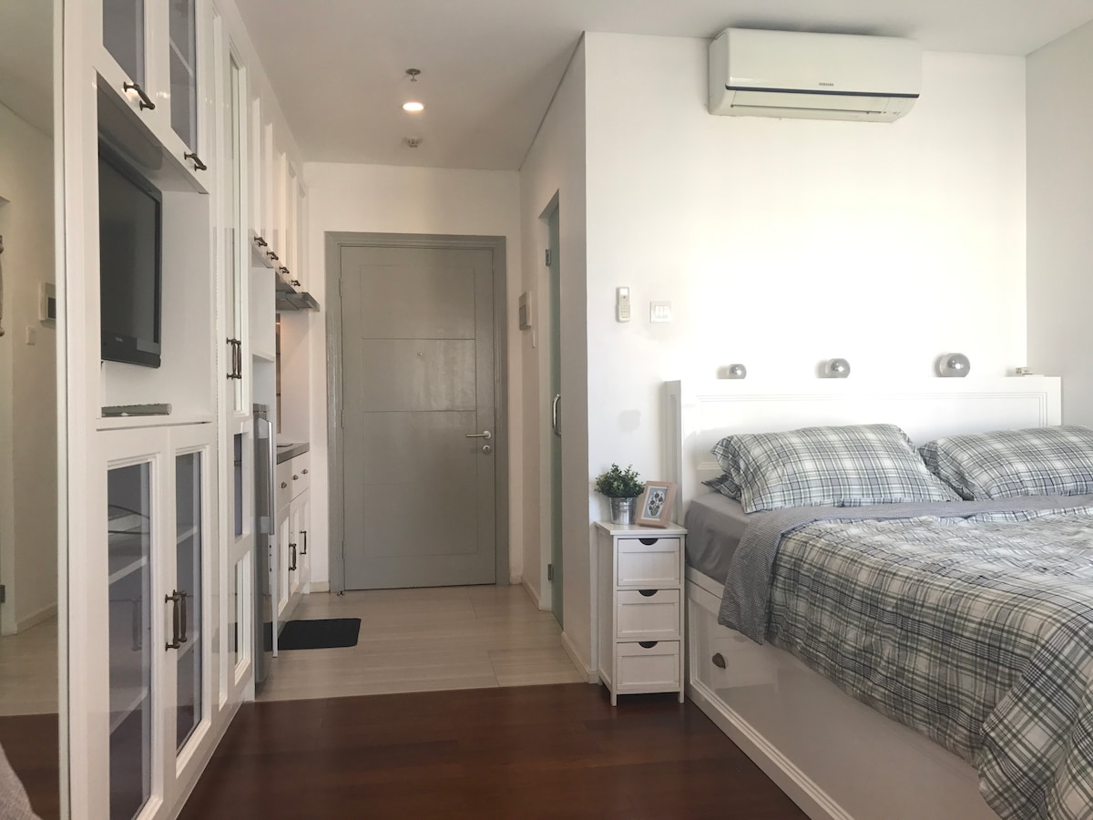 A well-organized studio space features a spacious bed adorned with a plaid duvet. Built-in storage solutions are visible, alongside a compact kitchen area. Natural light fills the room, complementing the contemporary decor. An air conditioning unit is mounted on the wall for climate control.