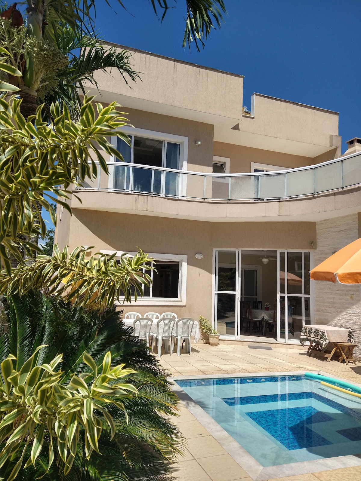The two-story house is depicted, featuring a modern exterior with large windows. A private pool with intricate tile work is seen at the forefront, surrounded by a patio area equipped with outdoor seating. Lush greenery frames the space, enhancing the tranquil environment.