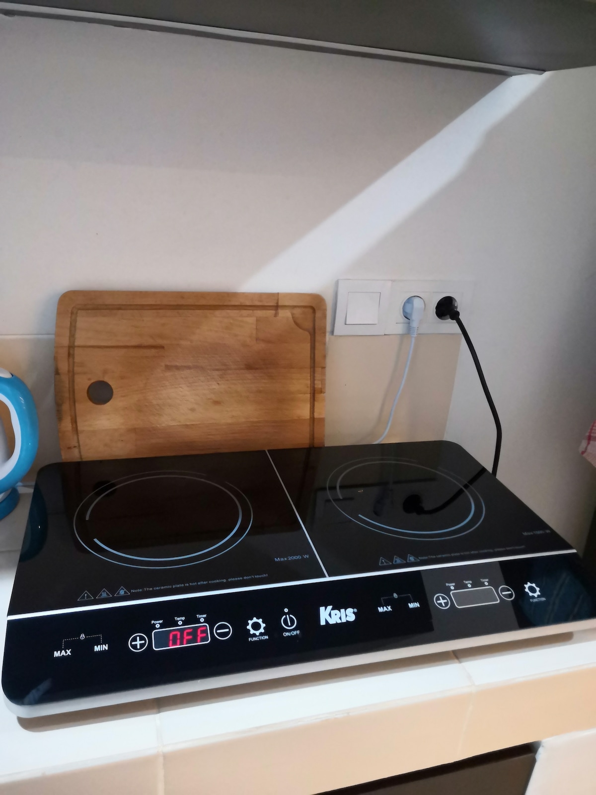 An electric cooking stove is displayed on a countertop, featuring two induction burners and control buttons. A wooden cutting board is positioned nearby, along with wall-mounted electrical outlets, providing a practical setup for meal preparation.