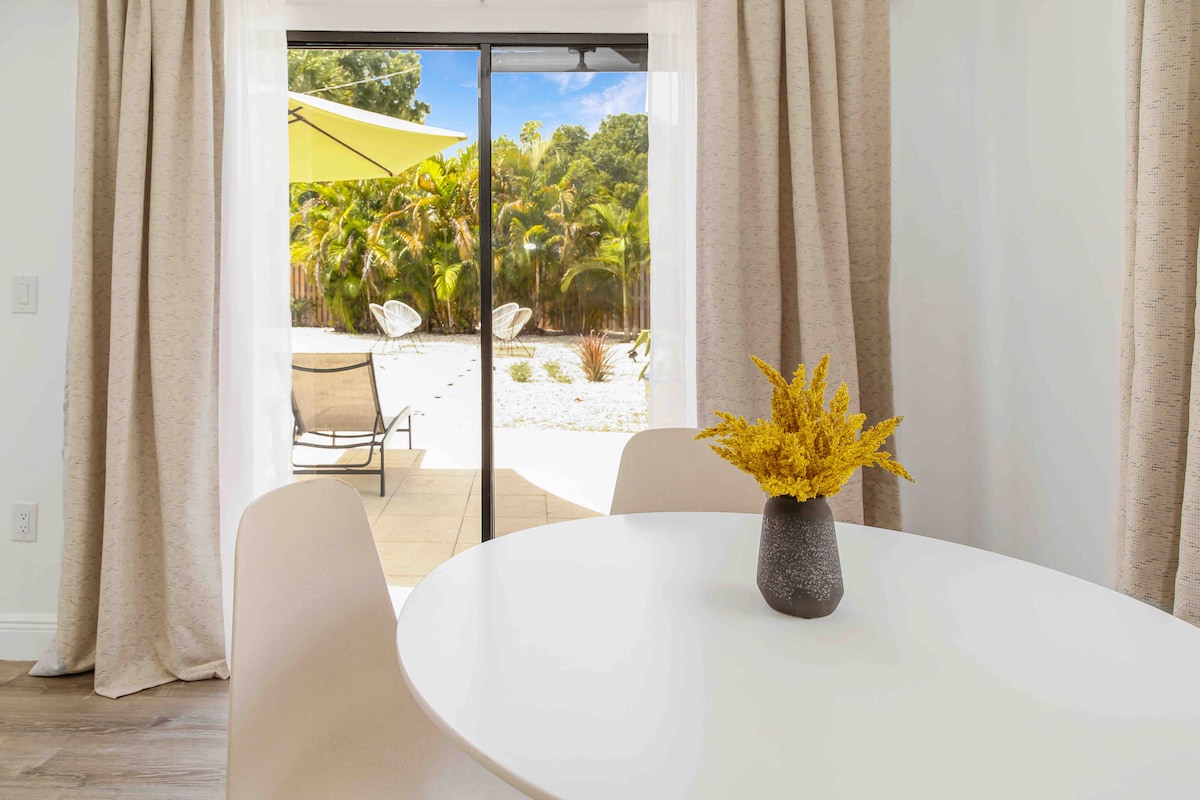 A dining area is visible, featuring a round white table set with a textured vase filled with yellow flowers. Light-colored curtains frame the sliding glass door that leads to a spacious patio and a landscaped backyard with palm trees.