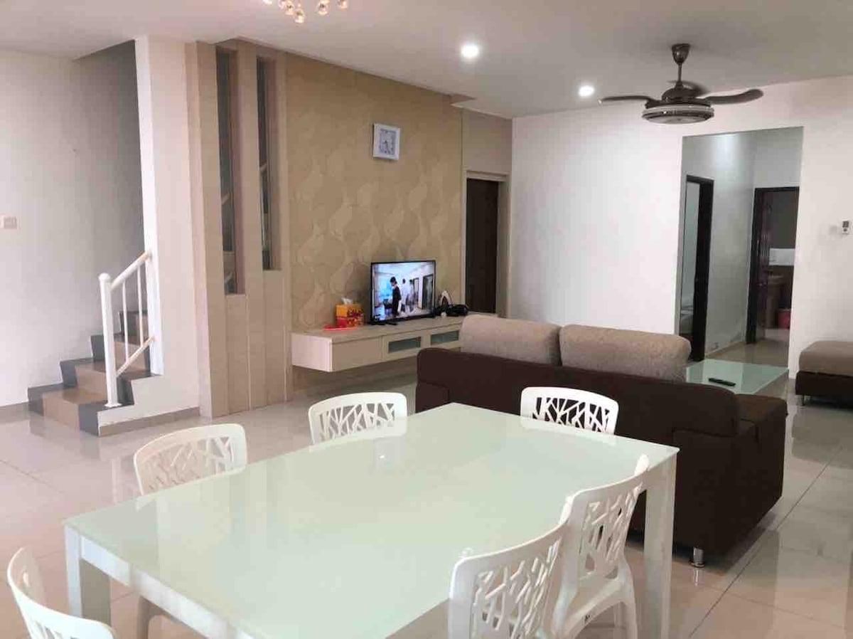 A modern living area featuring a comfortable sofa and a large television unit. A spacious dining table is surrounded by six white chairs. Soft lighting is present, and the space showcases an open layout with clean lines and neutral tones.