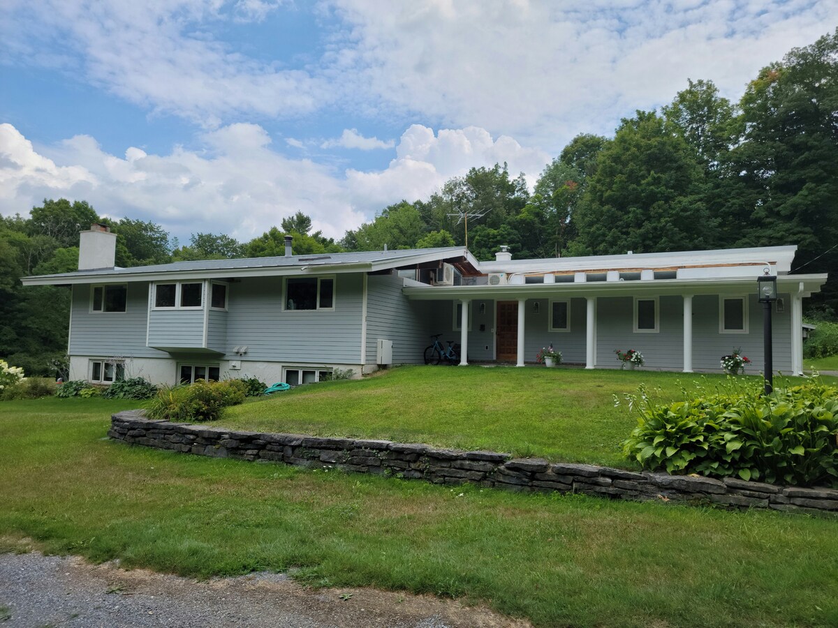 The exterior of a contemporary-style home is visible, surrounded by well-maintained green grass and a garden. The house features a combination of large windows and a broad roofline, set against a backdrop of trees and a blue sky with scattered clouds.