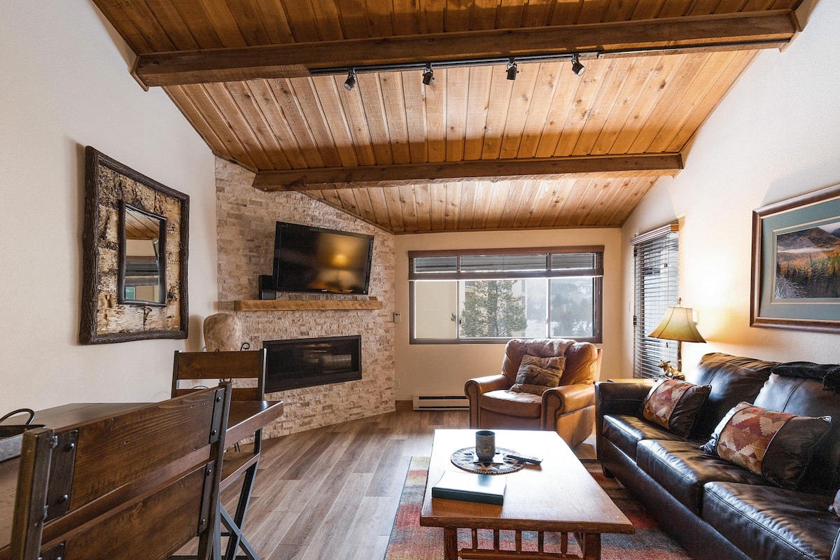 A living area features a vaulted wood ceiling and stone fireplace. A comfortable leather sofa and armchair provide seating. A wall-mounted television is above the fireplace, and a dining table with chairs is visible in the corner, enhancing the inviting space.