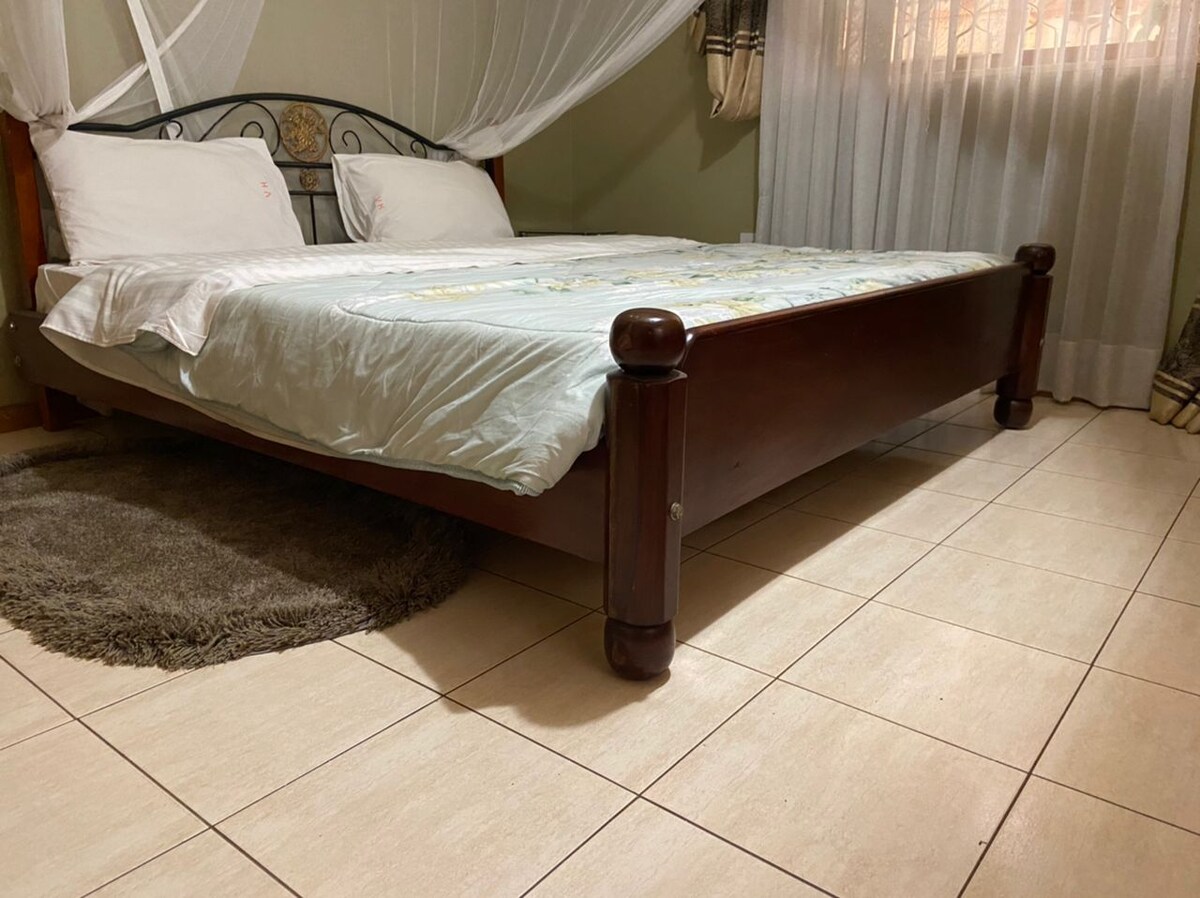 A spacious bed is positioned in the center of the room, draped with a light colored coverlet. The bed frame features rounded wooden posts. Soft natural light filters through sheer curtains, illuminating the tiled floor and a plush area rug beside the bed.