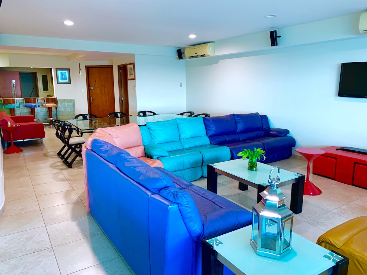 A spacious living area features a large sectional sofa in vibrant colors, along with a modern glass coffee table. An accent table adds additional seating options. The background reveals an open layout, including a dining set and a kitchen space, enhancing the communal feel.