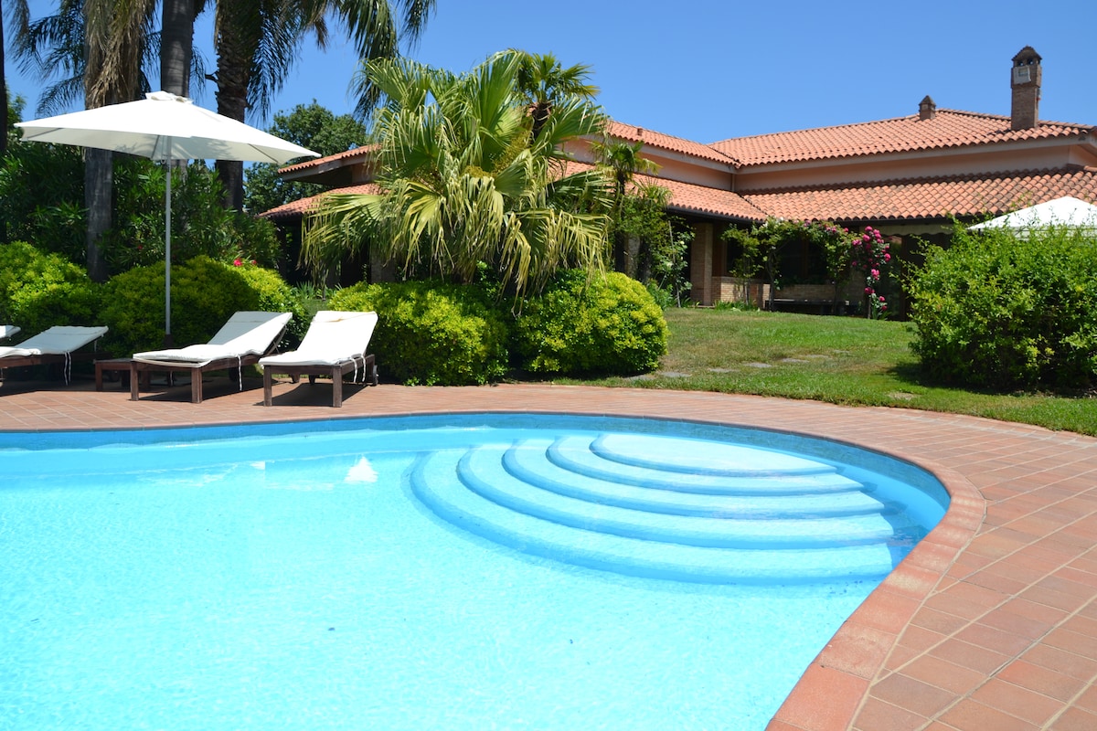 The inviting private pool is surrounded by lush greenery and neatly arranged deck chairs. A shaded parasol offers respite from the sun, while the villa's charming façade is visible in the background, enhanced by vibrant flowering plants and palm trees.