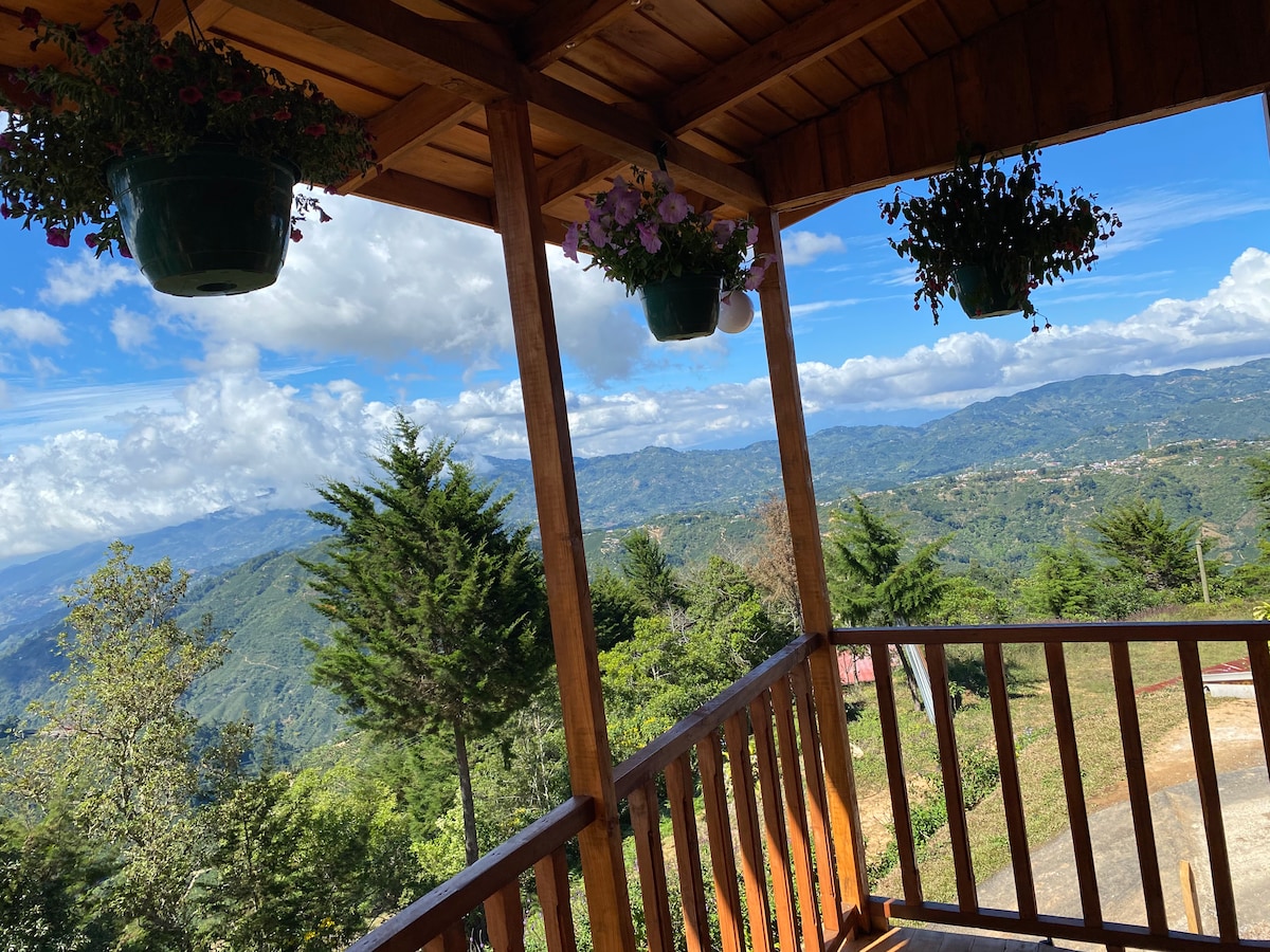 A wooden balcony offers a stunning view of the valley and surrounding mountains, framed by potted plants hanging from the overhead beams. The clear sky and scattered clouds enhance the natural beauty of the landscape, creating a peaceful outdoor space for relaxation.