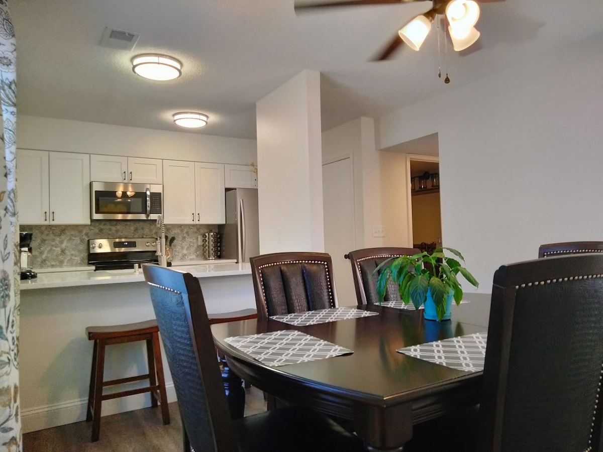 The dining area features a dark wooden table surrounded by six upholstered chairs. A potted plant adds a touch of greenery. In the background, a modern kitchen is visible, equipped with stainless steel appliances and white cabinetry.