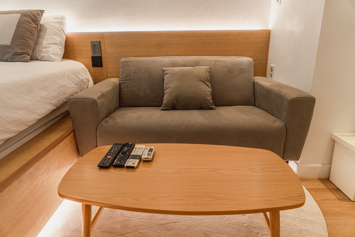 A simple seating area features a compact gray sofa positioned in front of a light wood coffee table. Multiple remote controls are arranged on the table. Soft lighting highlights the smooth surfaces, creating a clean and functional aesthetic.
