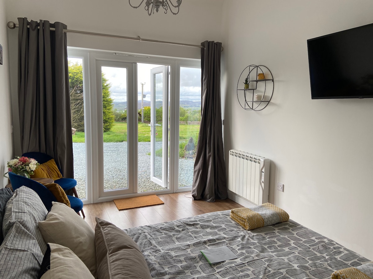 A bright living area features a comfortable seating arrangement, including a spacious sofa and cushions on the floor. Large glass doors open to reveal a gravel area and expansive views of the surrounding landscape. A wall-mounted TV and decorative shelf are also present.