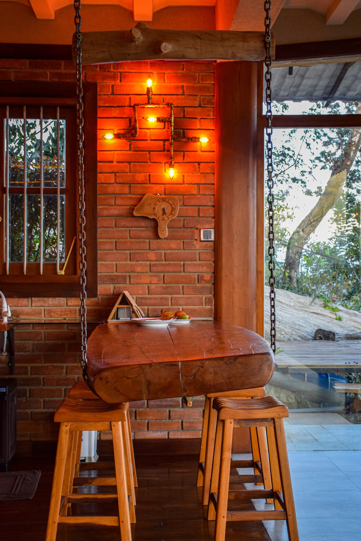 A rustic wooden swing table hangs from a sturdy beam, illuminated by warm Edison bulbs mounted on the brick wall. Two wooden stools are positioned beneath the table, contributing to a cozy and inviting dining area with large windows offering views of the surrounding nature.