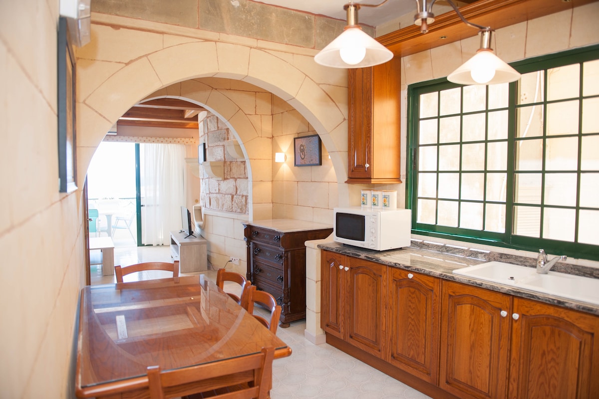 A kitchen area featuring wooden cabinetry and a countertop with a microwave. An archway leads to a dining space with a wooden table and chairs. Large windows provide natural light, showcasing the view beyond while soft lighting hangs above.