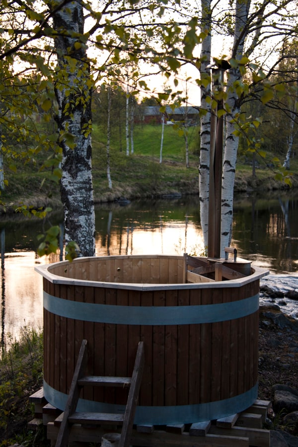 A wooden hot tub is positioned on a stone pier, surrounded by tall birch trees and reflecting water. The gentle glow of sunset illuminates the scene, enhancing the natural setting by Rickleån, with a calm river visible in the background.
