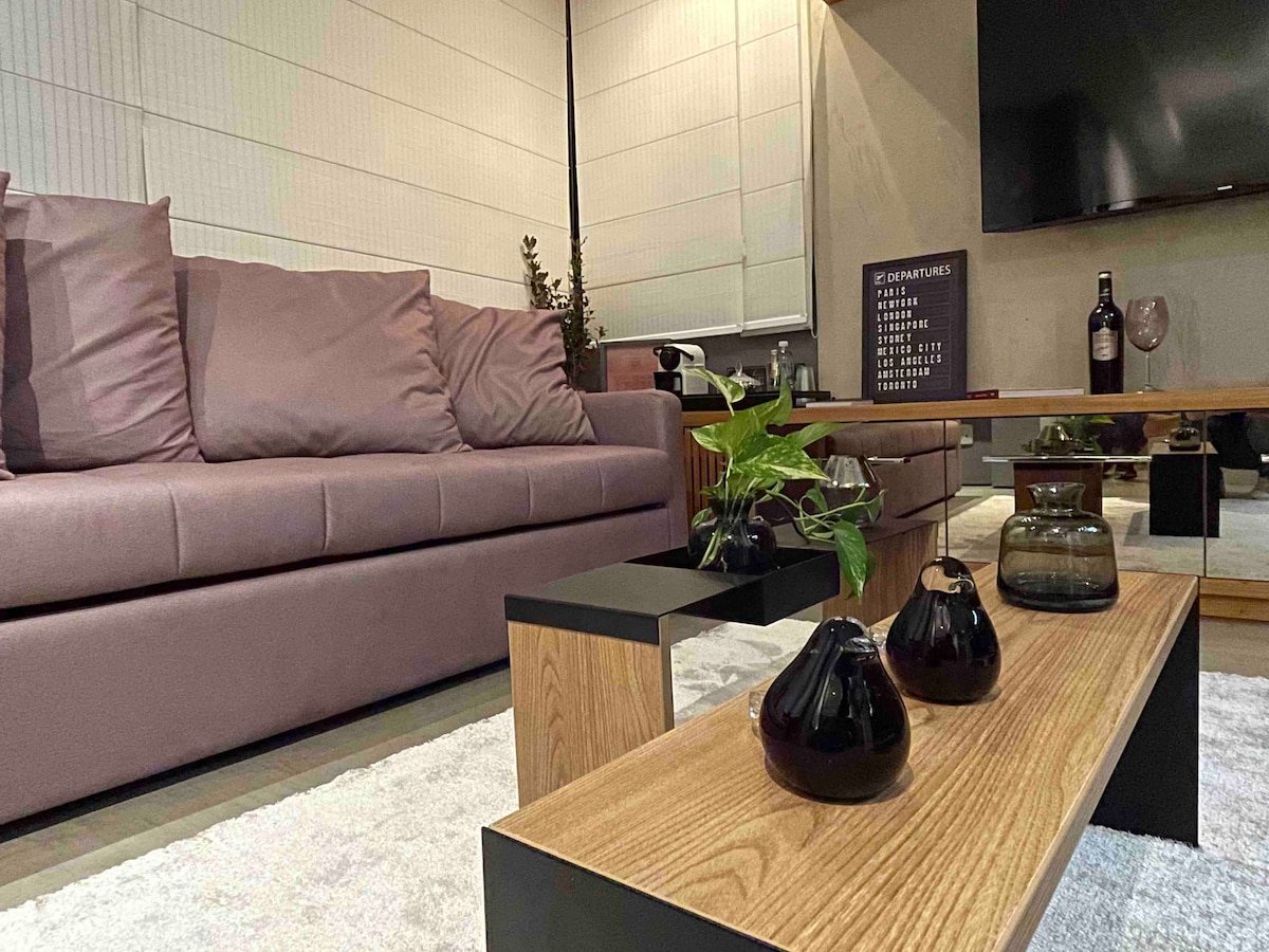 A stylish living area is presented with a plush mauve sofa, complemented by decorative cushions. A wooden coffee table holds a selection of dark glass vases and a small indoor plant. A flat-screen TV is mounted on the wall, enhancing the modern ambiance.