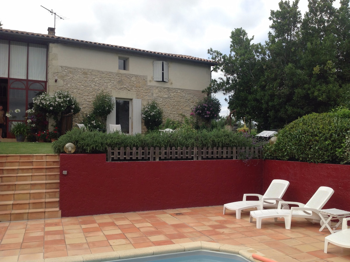 A view of the property from the swimming pool area, featuring a stone house with large windows and flowers lining the entrance. Several white loungers are placed around the pool, and neatly trimmed hedges create a boundary along the landscaped yard.