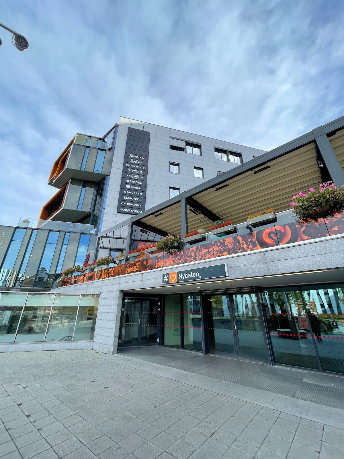 A modern building is seen featuring large glass windows and a prominent entrance labeled 'Nydalen.' The structure includes an outdoor terrace adorned with colorful flower arrangements, creating a welcoming atmosphere. The sky above is partly cloudy, providing natural light to the scene.