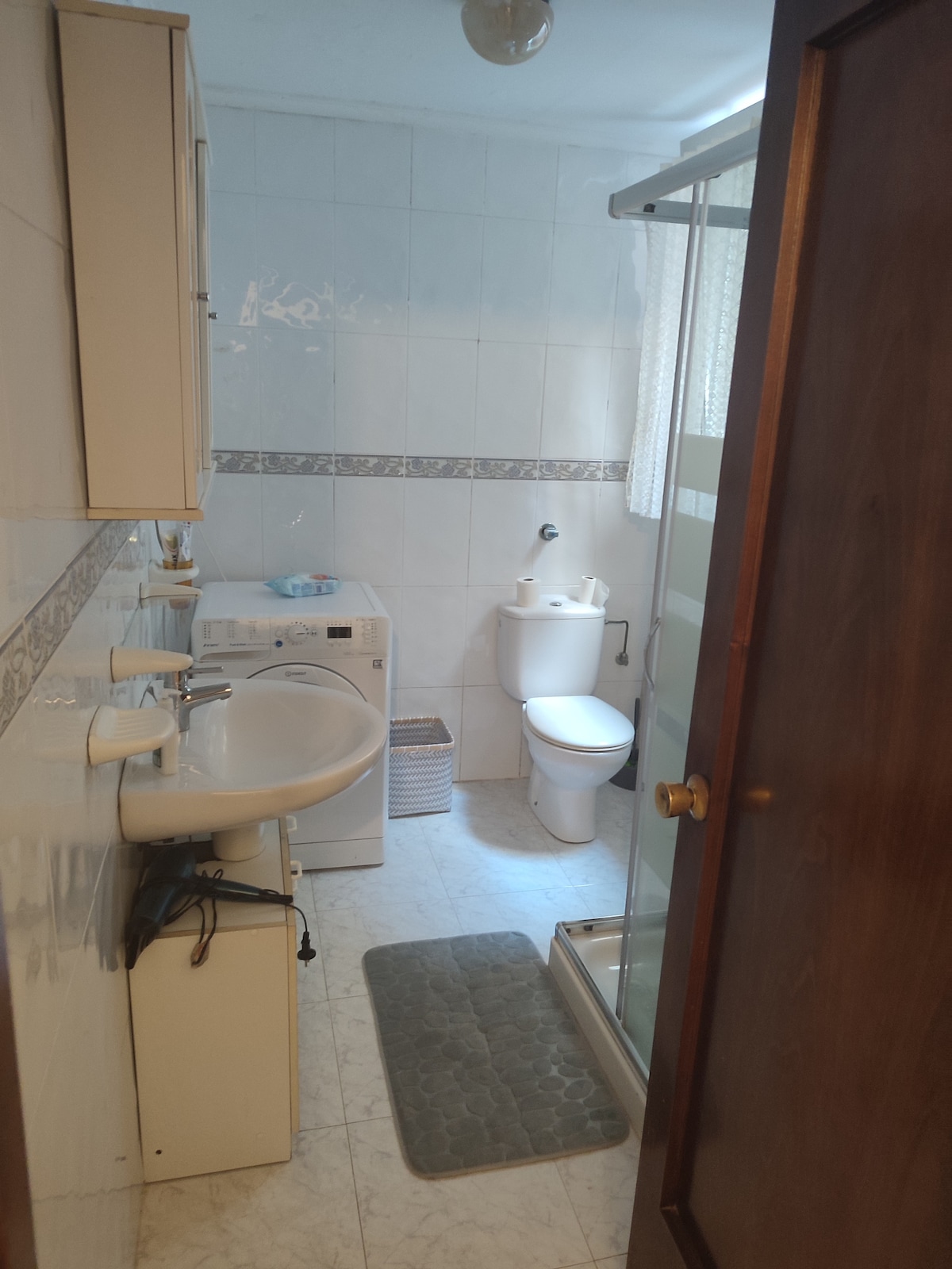A compact bathroom is visible, featuring a sink with storage underneath, a toilet, and a shower area with a glass door. Tiles line the walls, and soft natural light enters through a covered window. A washing machine is also present, enhancing the functionality of the space.
