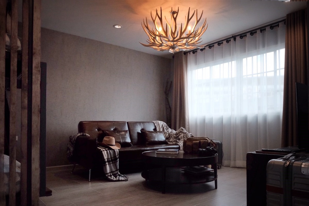 A spacious living area features a large, brown leather sofa adorned with a cozy throw and topped with a hat. A round coffee table sits centrally, surrounded by suitcases. Natural light filters through the sheer white curtains, illuminating the textured walls.