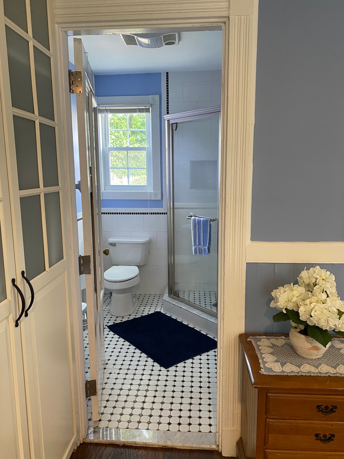 A well-lit bathroom is seen through a partially open door, featuring a walk-in shower, white toilet, and a window allowing natural light. A blue rug enhances the tiled floor, while a small vase of flowers decorates the nearby wooden dresser.