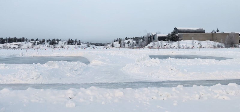 A snowy landscape stretches across the scene, with patches of icy surface partially visible. Colorful flags can be seen in the distance, marking a gathering area, while trees line the background against a gray sky. The scene captures the serene beauty of winter in Yellowknife.