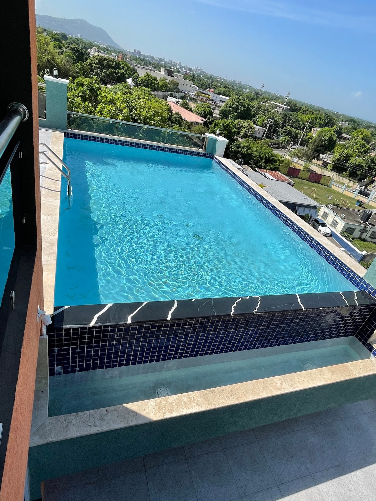 An outdoor pool is featured on a balcony, showcasing clear blue water with decorative tile accents. A view of greenery and distant hills can be seen in the background, offering an inviting space for relaxation.