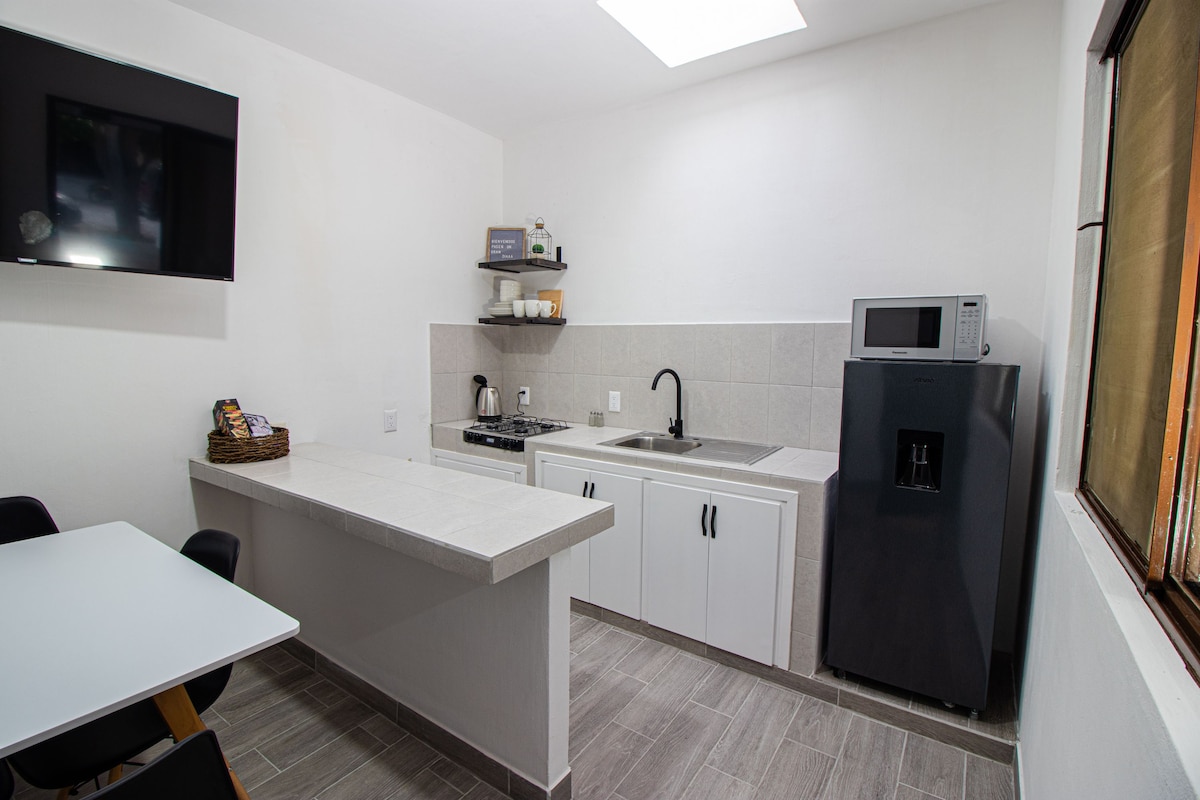 A modern kitchen showcases light-colored cabinetry and a large countertop. A stove, microwave, and refrigerator are arranged along the wall, with a sink located beneath a window. A dining table with black chairs is positioned nearby, providing space for gatherings.