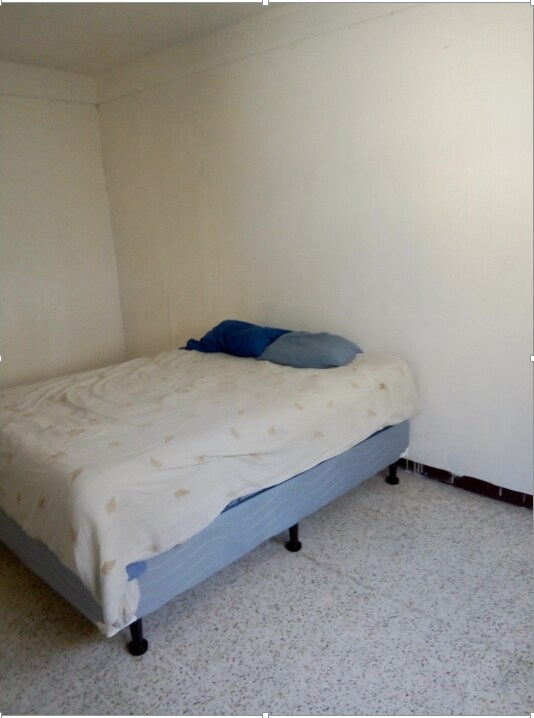 A simple sleeping area is presented, featuring a spacious bed covered with a light-colored blanket. Two pillows, one blue and one white, rest at the head of the bed. The walls are painted white, complementing the light floor tiles.