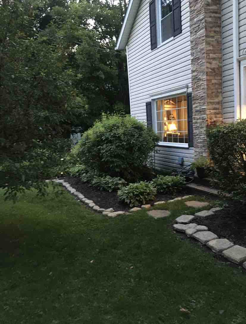 A landscaped garden area is visible alongside the home, featuring a stone pathway bordered by shrubs and greenery. Soft lighting from a window creates a welcoming glow, enhancing the natural surroundings and providing a sense of tranquility.