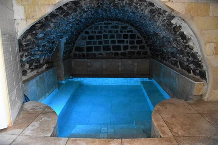 An indoor Turkish bath is enclosed by stone walls, featuring smooth, cool tiles and a serene blue pool of water. Soft lighting accentuates the arching ceiling, creating a tranquil environment perfect for relaxation.