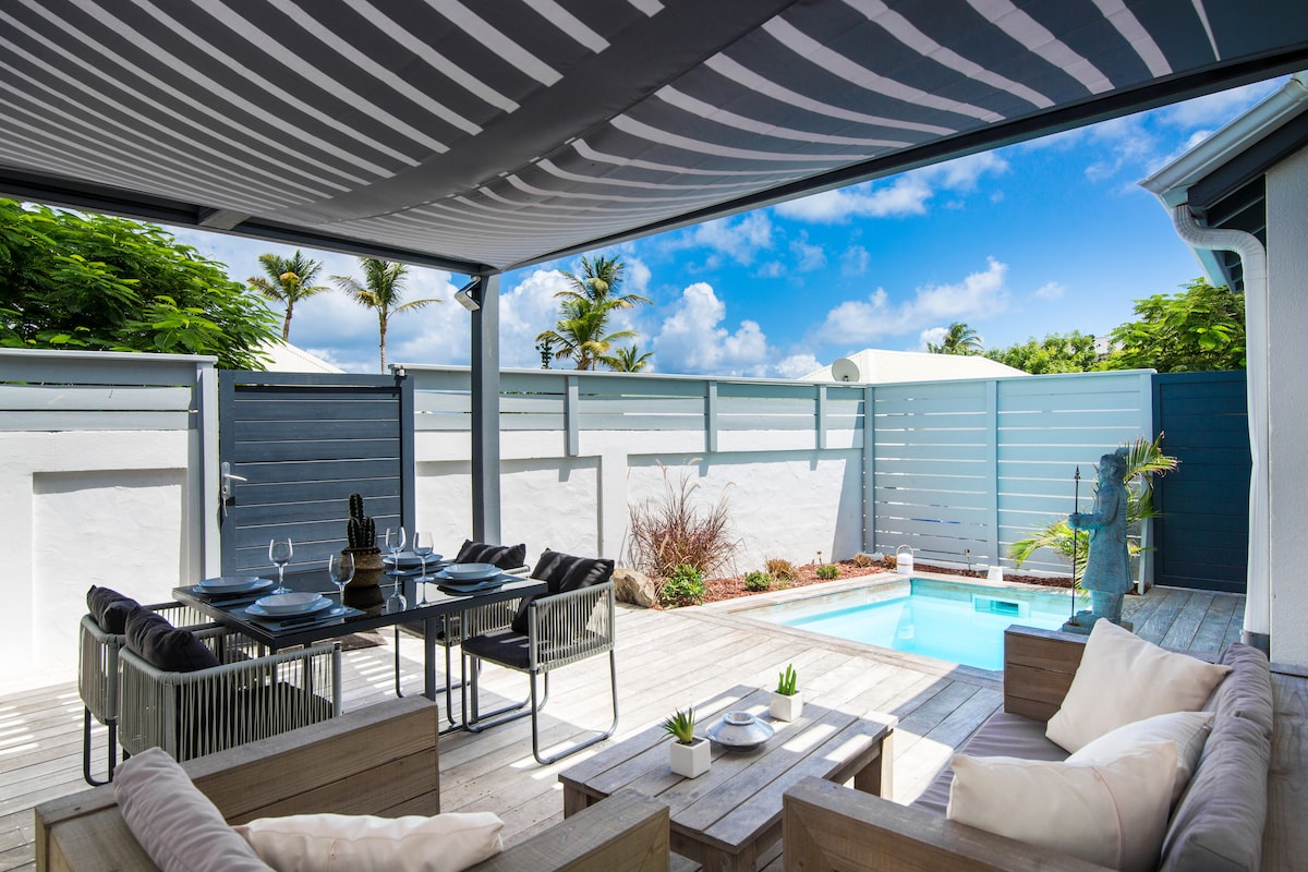 An outdoor seating area is showcased under a striped awning, featuring a wooden dining table set for six. A private pool is nestled nearby, surrounded by a wooden deck, while lush greenery and a low wall create a sense of privacy.