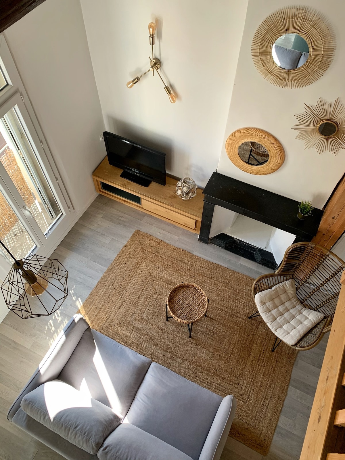 An overhead view of the living area displays a cozy sofa beside a natural fiber rug. A wooden media console supports a television, while decorative mirrors and a modern light fixture adorn the walls. A round side table and a rattan chair complete the inviting space.