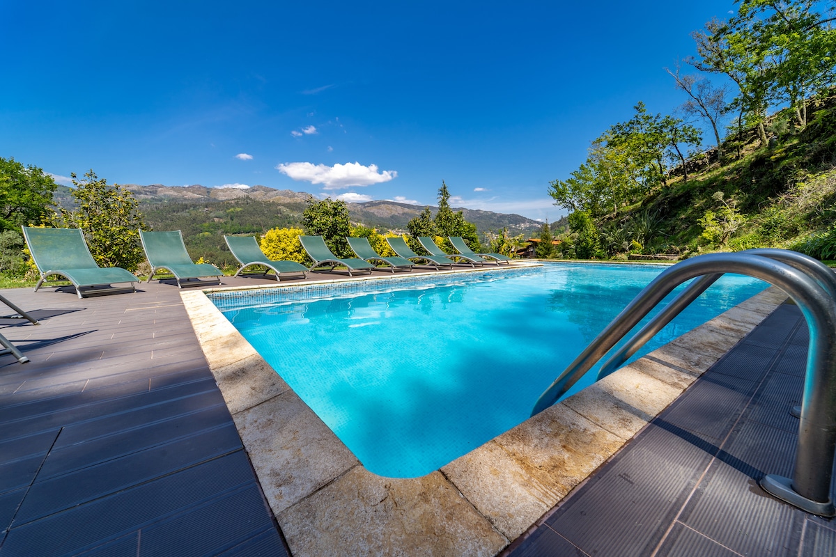 A private pool is framed by a spacious deck with numerous lounge chairs positioned for relaxation. Lush greenery and distant mountains create a serene backdrop, under a clear blue sky with a few scattered clouds.