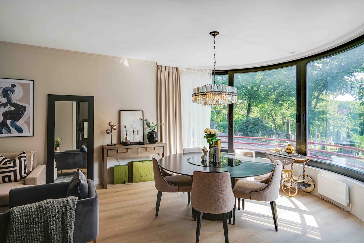 A stylish dining area features a round table surrounded by upholstered chairs. A crystal chandelier hangs overhead, with soft natural light filtering through large curved windows that offer a view of green foliage. A console table is visible, complemented by decorative elements.