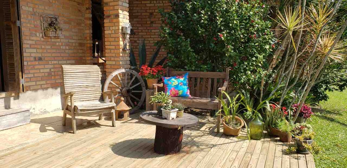An outdoor seating area is presented with wooden chairs and a small round table at the center. Vibrant potted plants surround the space, complementing the greenery in the background. A decorative wheel adds a rustic touch to the inviting area.