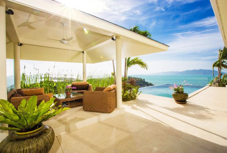 An outdoor seating area is presented with comfortable wicker furniture, surrounded by lush greenery. The view extends over the infinity pool to the sea, with a bright sky enhancing the tranquil atmosphere. Sunlight casts a warm glow on the space.