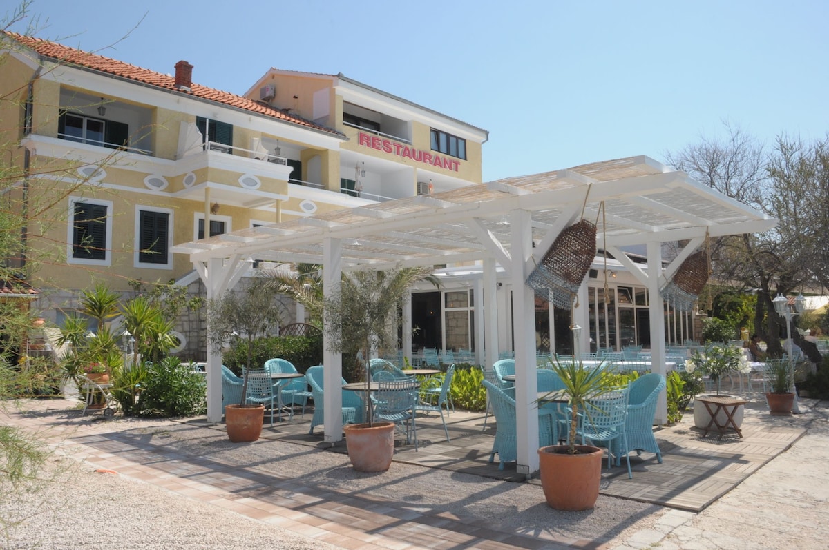 An outdoor dining area features a white wooden pergola adorned with hanging wicker baskets. Light blue chairs surround round tables, positioned amid an array of potted plants. A building with a restaurant sign is visible in the background, with clear blue skies above.