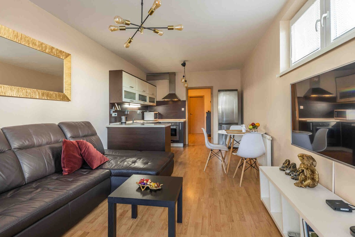 A modern living space features a dark leather sofa with a decorative pillow, paired with a low black table. The kitchen area displays contemporary cabinetry and appliances. A dining table set is positioned nearby, with a fruit bowl as a centerpiece. Natural light enters through large windows.