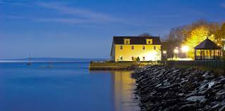A charming cottage is positioned along the shoreline, illuminated by soft lighting. The calm waters of the bay reflect the evening sky, while nearby rocks create a textured contrast. Trees provide natural framing, enhancing the serene atmosphere of the coastal setting.