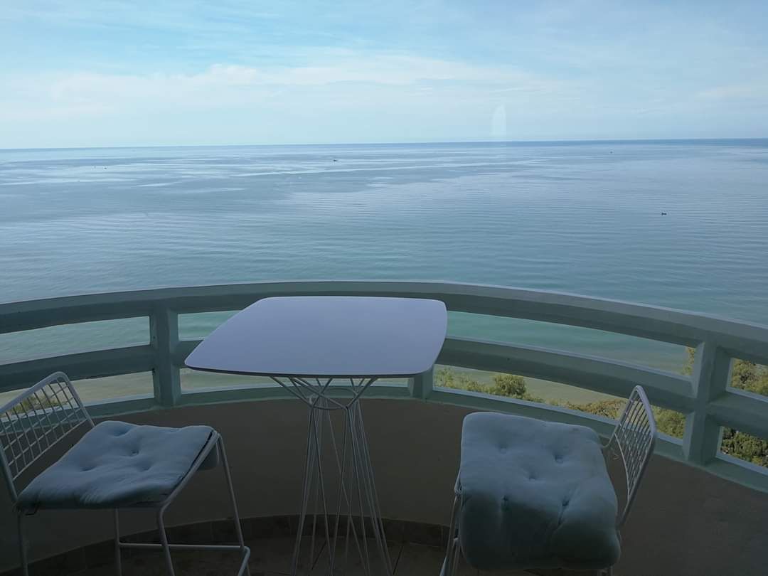 A small outdoor balcony features a round table and two modern chairs, all positioned to offer expansive views of the calm ocean waters. The horizon blends seamlessly with the sky, creating a serene coastal atmosphere.